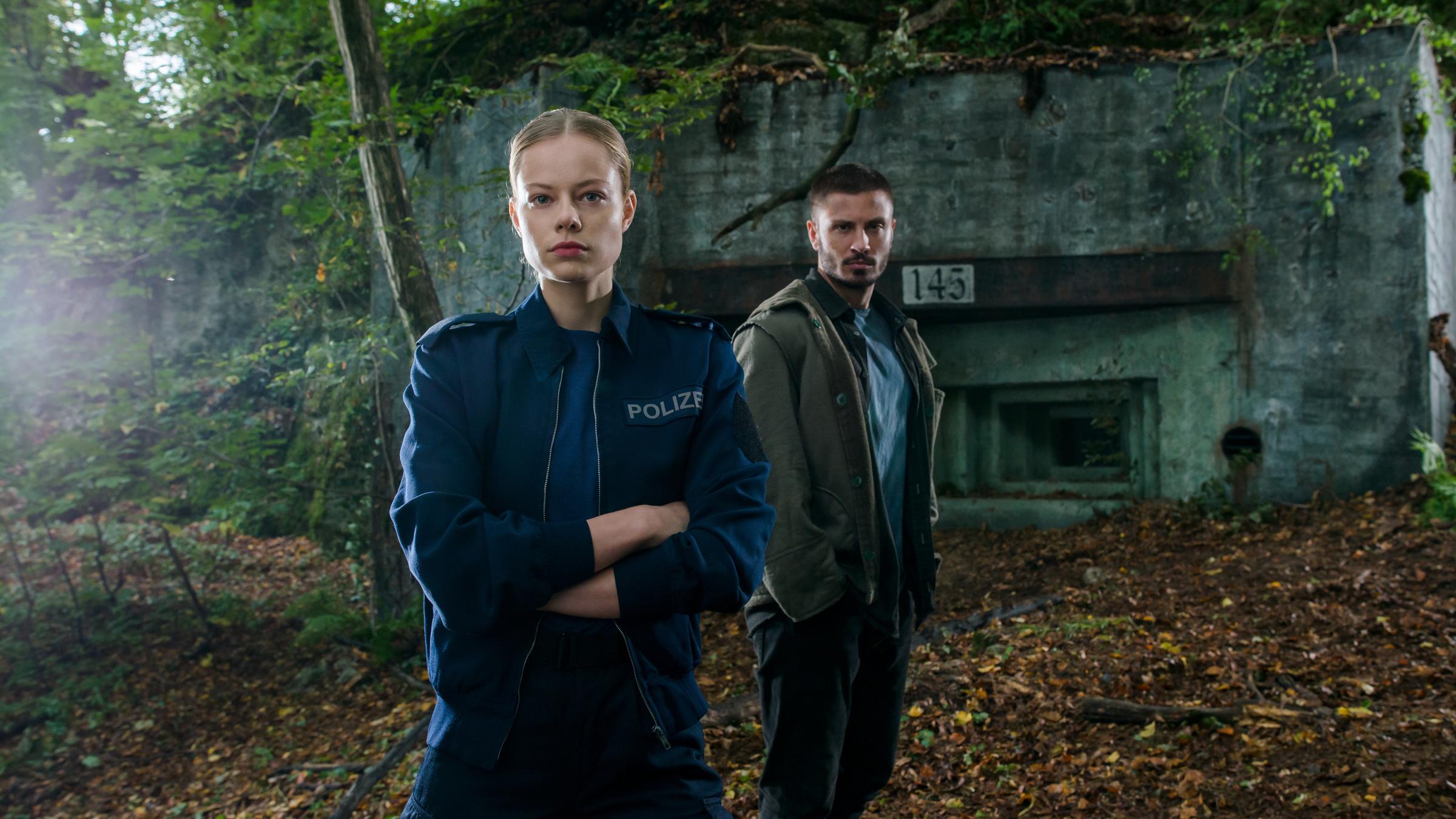 "Westwall - Kapitel I": Julia (Emma Bading) und Nick (Jannik Schümann) stehen vor einer alten Bunkeranlage im Wald. Julia hat die Arme vor der Brust verschränkt, Nick steht etwas hinter ihr, die Hände in den Hosentaschen. Beide schauen in die Kamera.