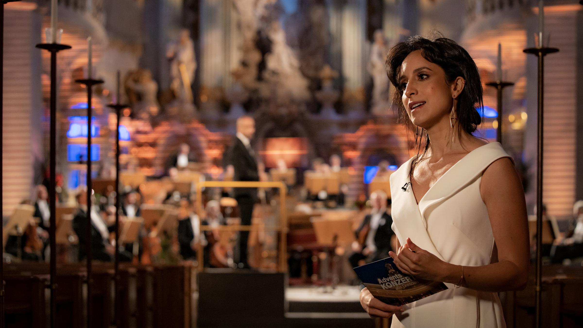 "Adventskonzert aus Dresden": Stephanie Stumph steht vor einer Bühne, auf der Musiker unscharf zu erkennen sind.