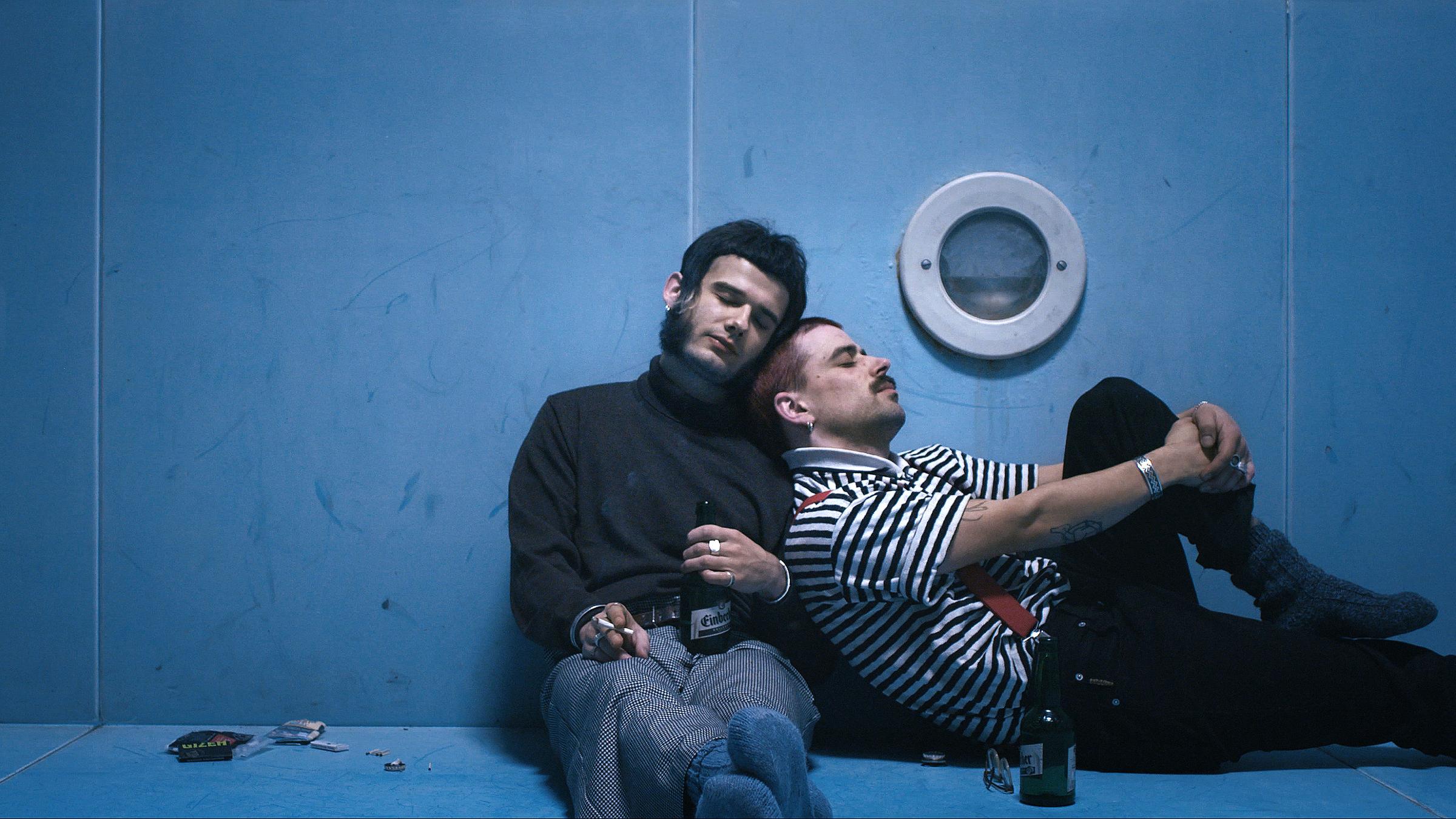 "Ab 18! – Fehler und Irritation": Zwei junge Männer sitzen in dämmrigem Licht auf dem Boden eines Swimming Pools, in dem kein Wasser ist. Der links sitzende mit Rollkragenpullover lehnt an die Mauer des Pools. Er hält eine brennende Zigarette in der – vom Betrachter aus gesehen – linken und eine Bierflasche in der rechten Hand. An ihn lehnt sich der zweite Mann mit Schnurrbart und blau gestreiftem T-Shirt, der im 90°-Winkel zu ihm sitzt. Er hat ein Bein angezogen. Beide haben die Augen geschlossen.