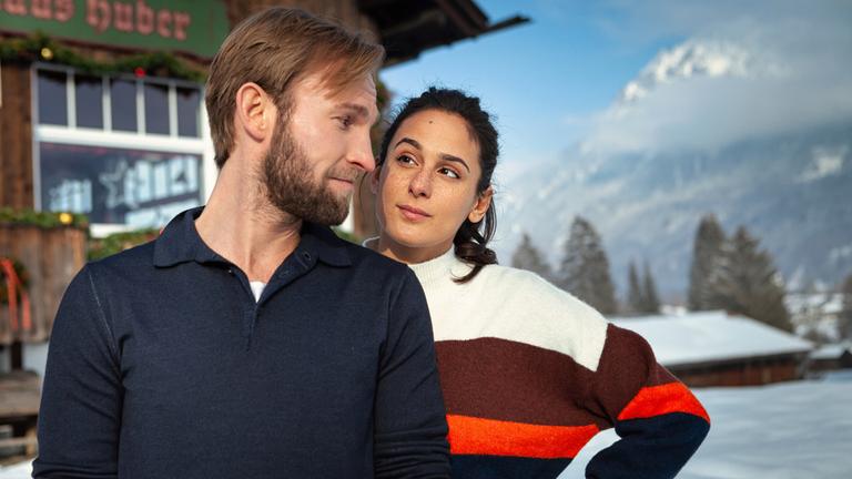 "Herzkino - Alice im Weihnachtsland": Jakob (Jochen Matschke) mit einem blauen Pullover und eine  Alice (Aybi Era) mit einem gestreiften Pullover stehen dicht nebeneinander vor einem Holzhaus. Er blickt sie herausfordernd an, sie hat skeptisch eine Augenbraue hochgezogen. Es liegt Schnee und die Sonne scheint.