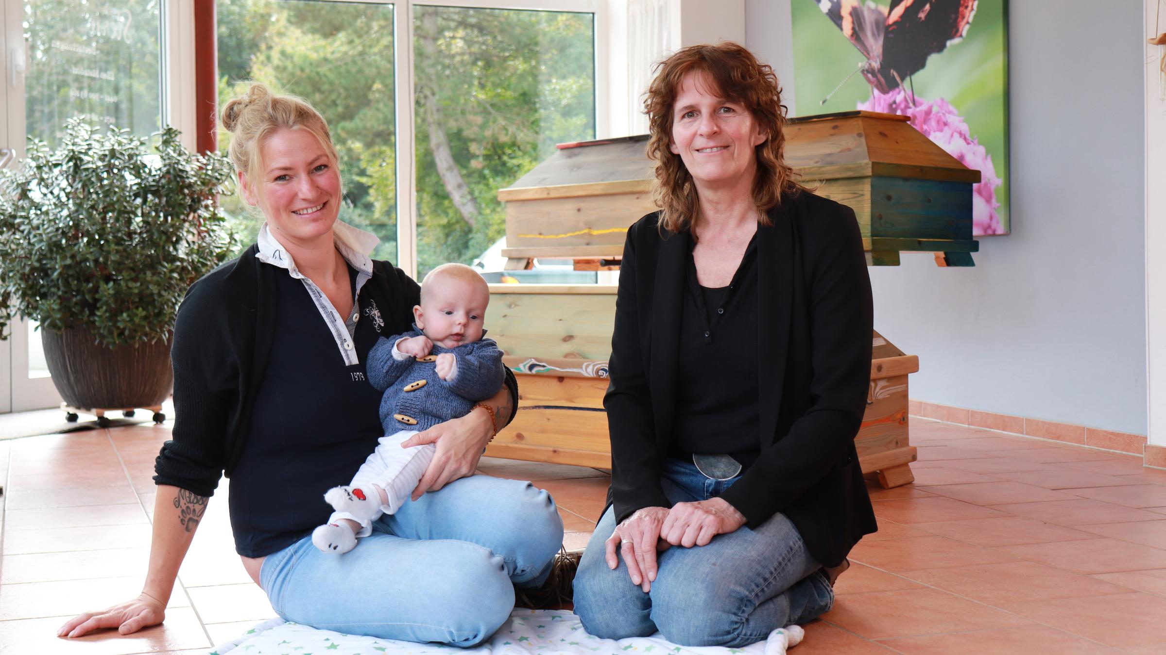 "37° Vom Anfang und Ende des Lebens - Arbeiten als Hebamme und Bestatterin": Ellen Matzdorf und Laetitia Lübcke mit Sohn Levi sitzen auf einer Decke vor zwei Ausstellungssärgen.