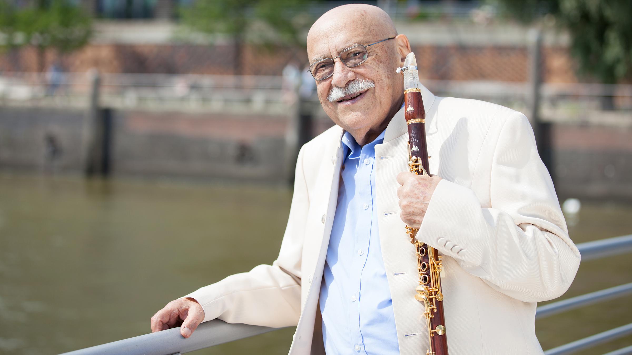 "Happy Birthday, Giora Feidman!": Giora Feidman steht auf einer Brücke, ein Arm liegt auf dem Brückengeländer. In der anderen Hand hält er die Klarinette.