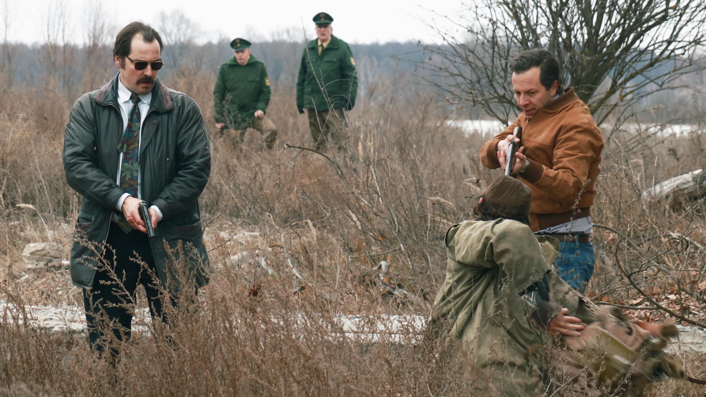 "Freies Land": Bach (Felix Kramer) und Stein (Trystan Pütter) stehen im Gestrüpp und richten ihre Waffen gegen einen Wilderer (Ben Hartmann), der vor ihnen auf dem Boden kniet. Im Hintergrund sind Polizisten zu sehen, die das Geschehen beobachten.