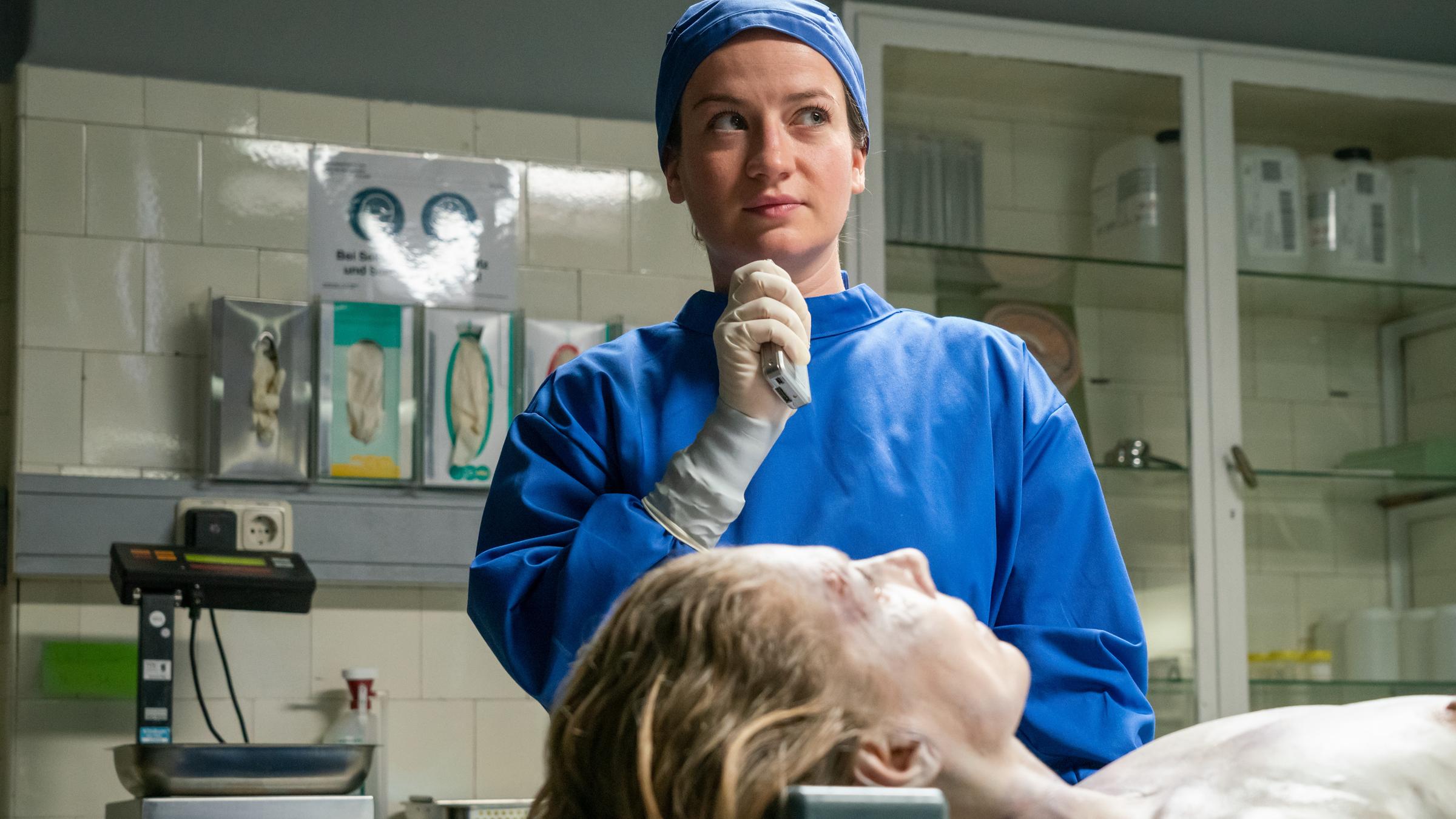 "Theresa Wolff - Home Sweet Home": Theresa Wolff (Nina Gummich) steht neben der aufgebahrten Toten (Antje Rau) am Obduktionstisch und schaut in die Ferne. Sie trägt einen blauen Kittel und eine blaue Haube.