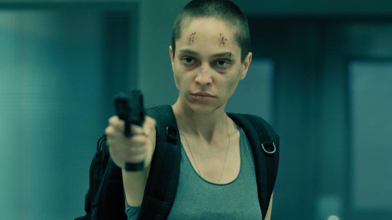 "Box 21 - Nicht aufgeben": Lidia Amariei (Ilinca Neacșu) zielt mit einer Waffe auf ihr Gegenüber. Ihr Kopf ist rasiert und im Gesicht hat sie mehrere Wunden und blaue Flecken. Ihr Gesichtsausdruck ist kalt und fokussiert.