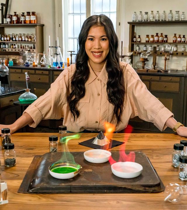 "Terra X: - Wunderwelt Chemie - Die Bausteine der Natur": Mai Thi Nguyen-Kim lehnt in einem alten Labor an einem Tisch. Vor ihr verschiedene Schalen mit chemischen Stoffen, die in grün, rot und gelb leuchten.