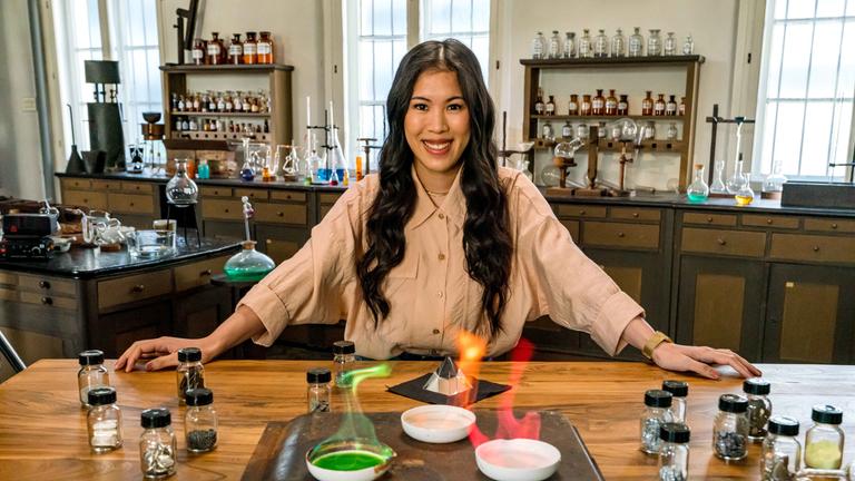 "Terra X: - Wunderwelt Chemie - Die Bausteine der Natur": Mai Thi Nguyen-Kim lehnt in einem alten Labor an einem Tisch. Vor ihr verschiedene Schalen mit chemischen Stoffen, die in grün, rot und gelb leuchten.