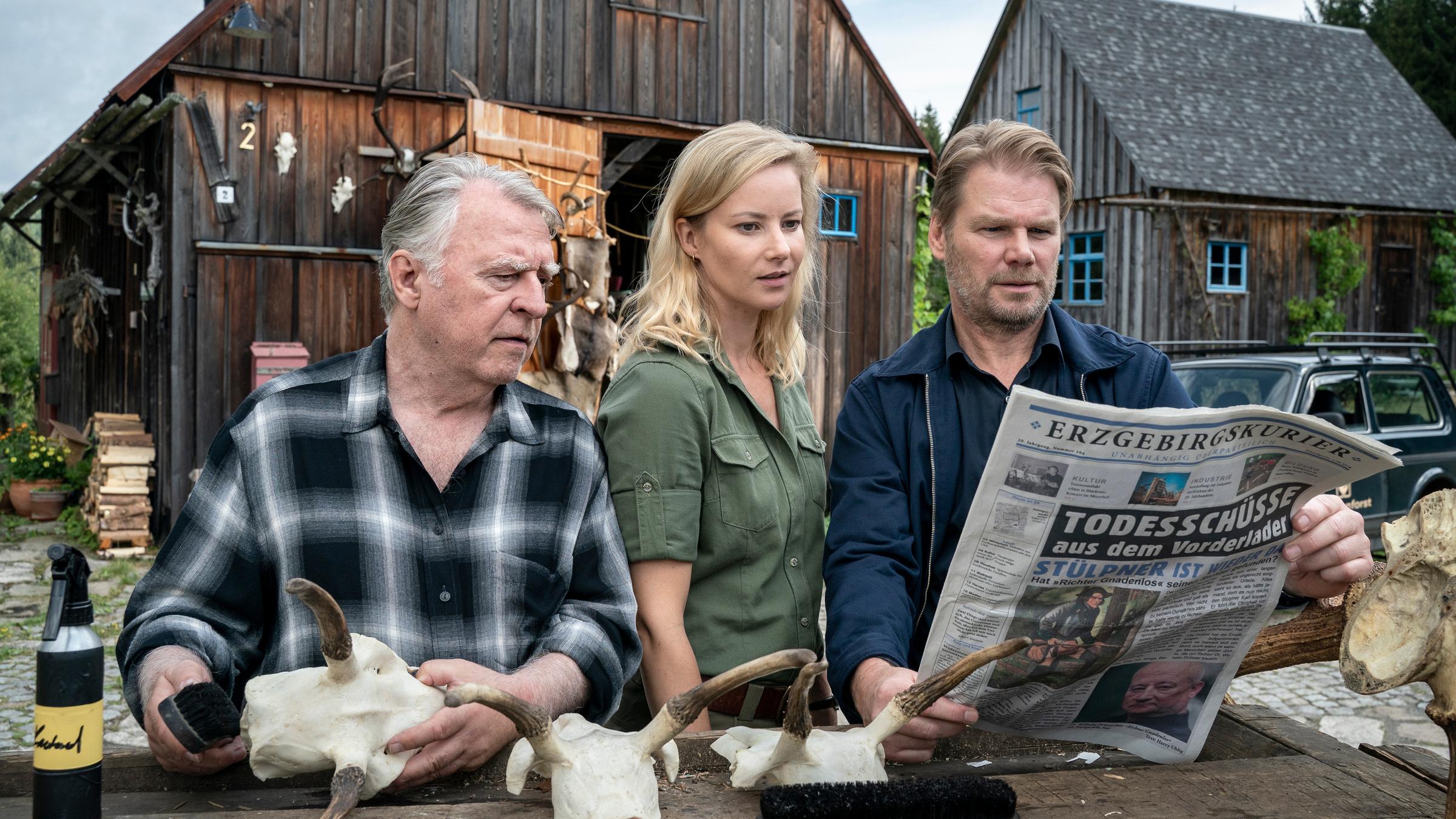 "Erzgebirgskrimi - Der letzte Bissen": Außenaufnahme. Georg Bergelt (Andreas Schmidt-Schaller), Saskia Bergelt (Teresa Weißbach) und Robert Winkler (Kai Scheve) stehen im Hof des Forstamtes an einem Tisch mit Geweihen und schauen in eine Zeitung, deren Schlagzeile lautet: "Todesschuss mit Vorderlader".