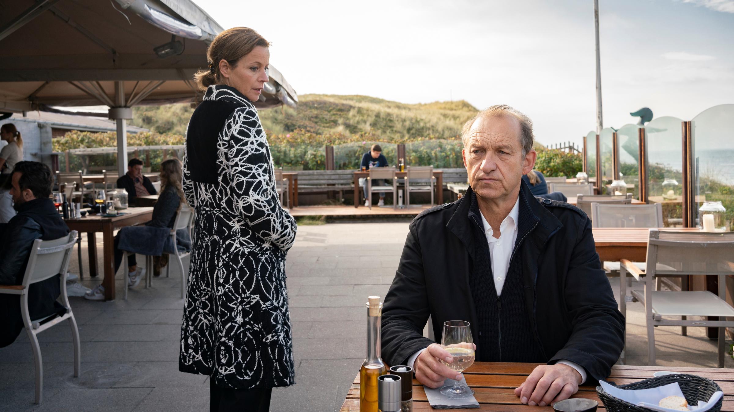 "Nord Nord Mord - Sievers und der schwarze Engel": Carl Sievers (Peter Heinrich Brix) sitzt auf einer Restaurantterrasse an einem braunen Holztisch. Er hat ein Glas Wasser in der rechten Hand und schaut nachdenklich nach vorn. Schräg links hinter ihm im Gang zwischen den Tischen - und in Carl Sievers Rücken - steht eine Frau, Hanne Wegener (Jule Ronstedt), in einem schwarzen Mantel mit weißem Muster, die sich leicht nach vorne dreht und Carl Sievers von hinten anschaut.