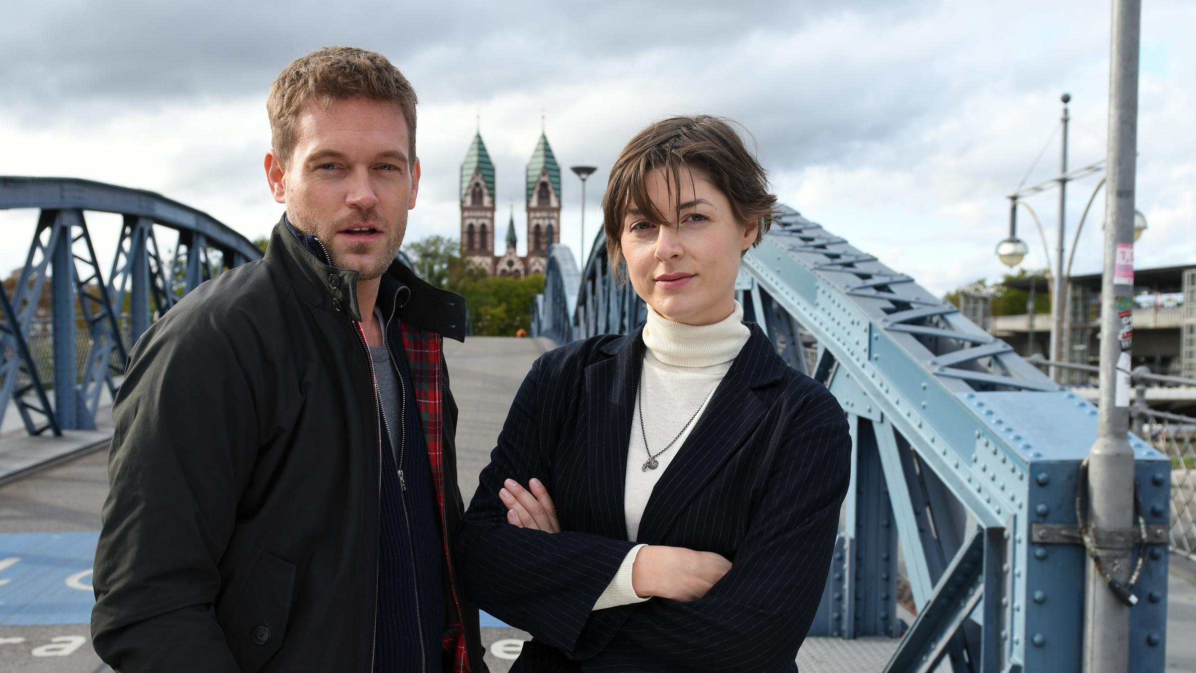 "Breisgau - Bullenstall": Tanja Wilken (Katharina Nesytowa) mit verschränkten Armen und Dennis Danzeisen (Joscha Kiefer) stehen vor einer Brücke in Freiburg und blicken gerade aus. Im Hintergrund steht eine Kirche mit zwei Kirchtürmen.