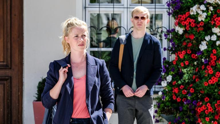 "Marie fängt Feuer - Schattenhaft": Marie Reiter (Christine Eixenberger) steht mit einer Tasche über die Schulter gelehnt neben einem Blumenschmuck, während Mark Gruber (David Zimmerschied) im Hintergrund vor dem Gebäude der Bürgersprechstunde steht.