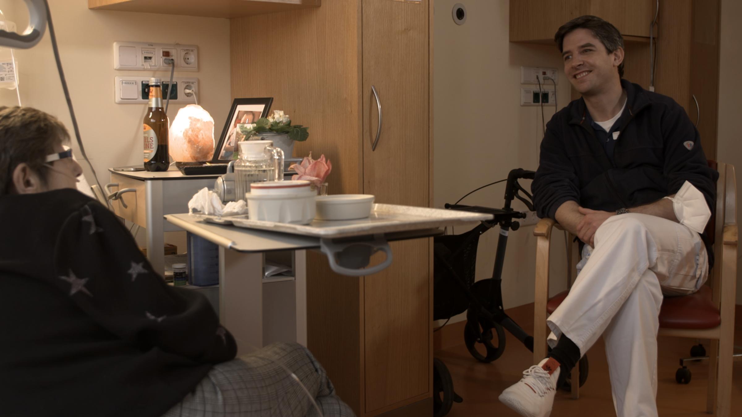 "37° Die letzten guten Tage - Wie Palliativärzt*innen helfen": In einem Krankenhauszimmer sitzt Palliativarzt Dr. Philipp von Trott einer Patientin gegenüber und lächelt sie an.