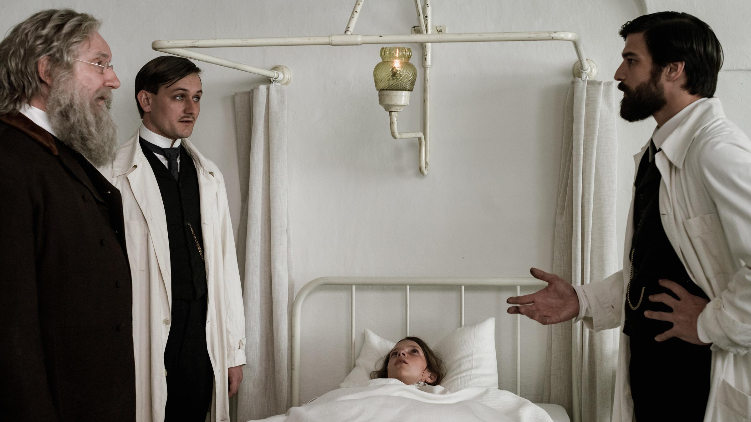 "Freud - Trauma": Freud (Robert Finster) steht vor Leopold von Schönberg (Lukas Watzl) und Prof. Dr. Theodor Meynert (Rainer Bock) und blickt sie ernst an. Zwischen ihnen liegt Clara von Schönfeld (Zuzana Zvonícková) im Krankenbett.