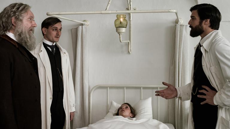 "Freud - Trauma": Freud (Robert Finster) steht vor Leopold von Schönberg (Lukas Watzl) und Prof. Dr. Theodor Meynert (Rainer Bock) und blickt sie ernst an. Zwischen ihnen liegt Clara von Schönfeld (Zuzana Zvonícková) im Krankenbett.