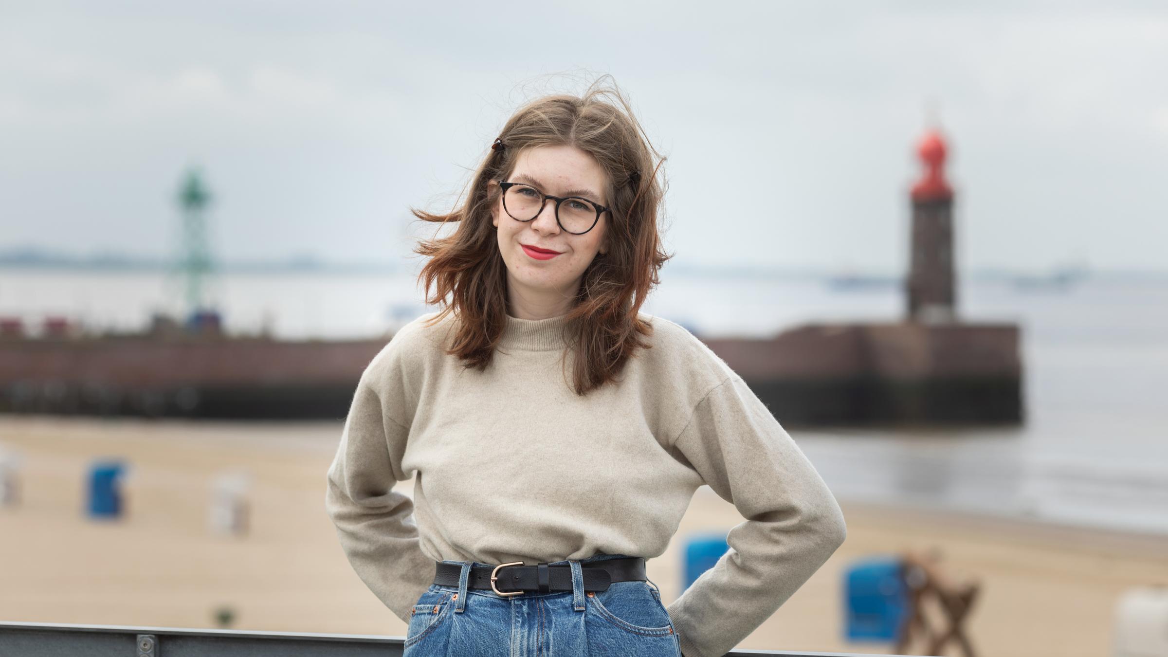 "Einfach Mensch": Jennifer Wrona steht vor Geländer. Im Hintergrund Strand und Leuchtturm.