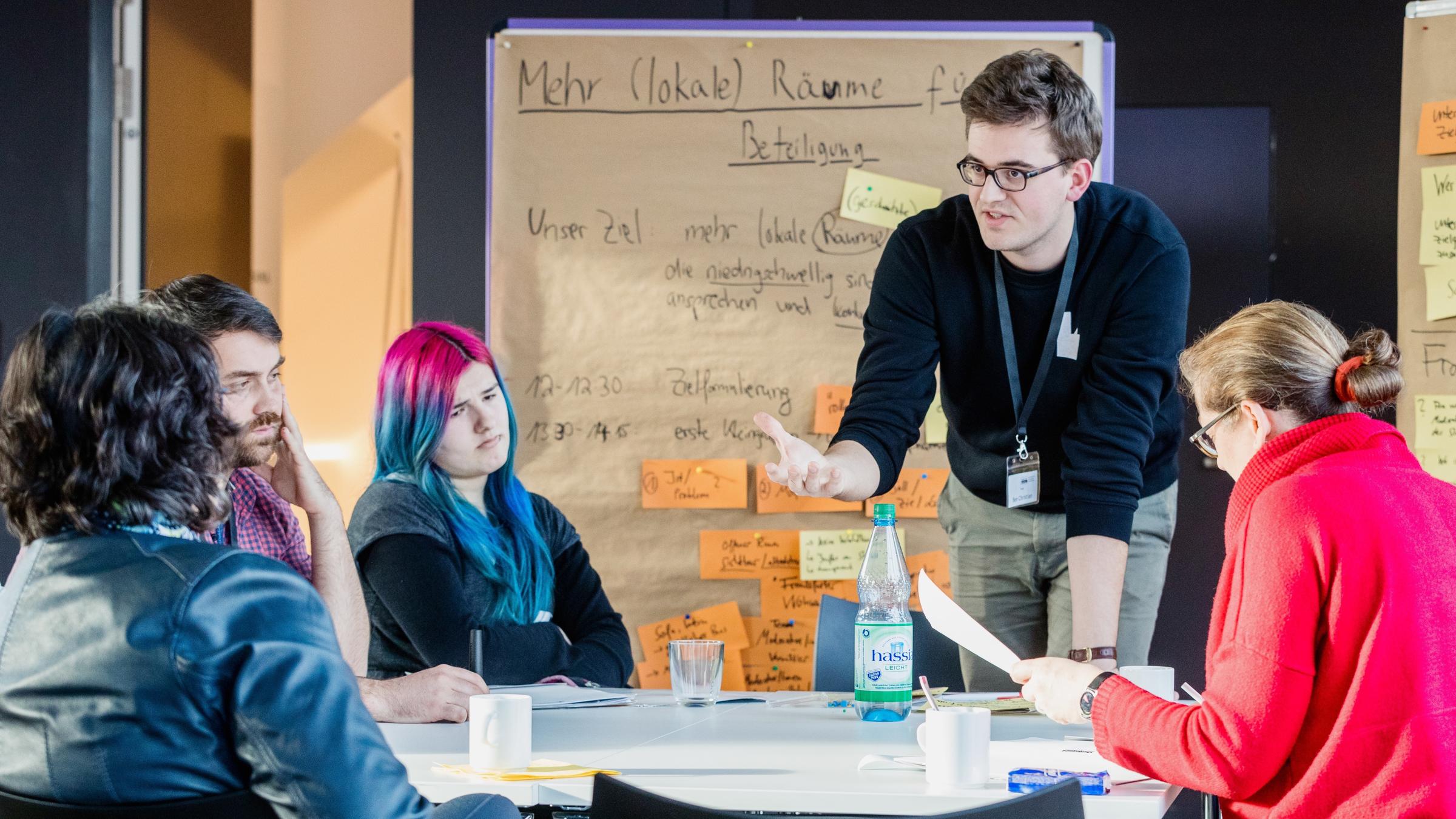 "plan b: Wir machen Politik - Mitreden, mitgestalten, mitentscheiden": Eine Gruppe von zwei Männern und zwei Frauen sitzen an einem Tisch hören auf einen Mann, der vor ihnen steht und redet. Im Hintergrund eine beschriftete Tafel mit orangen und gelben Notizzetteln.