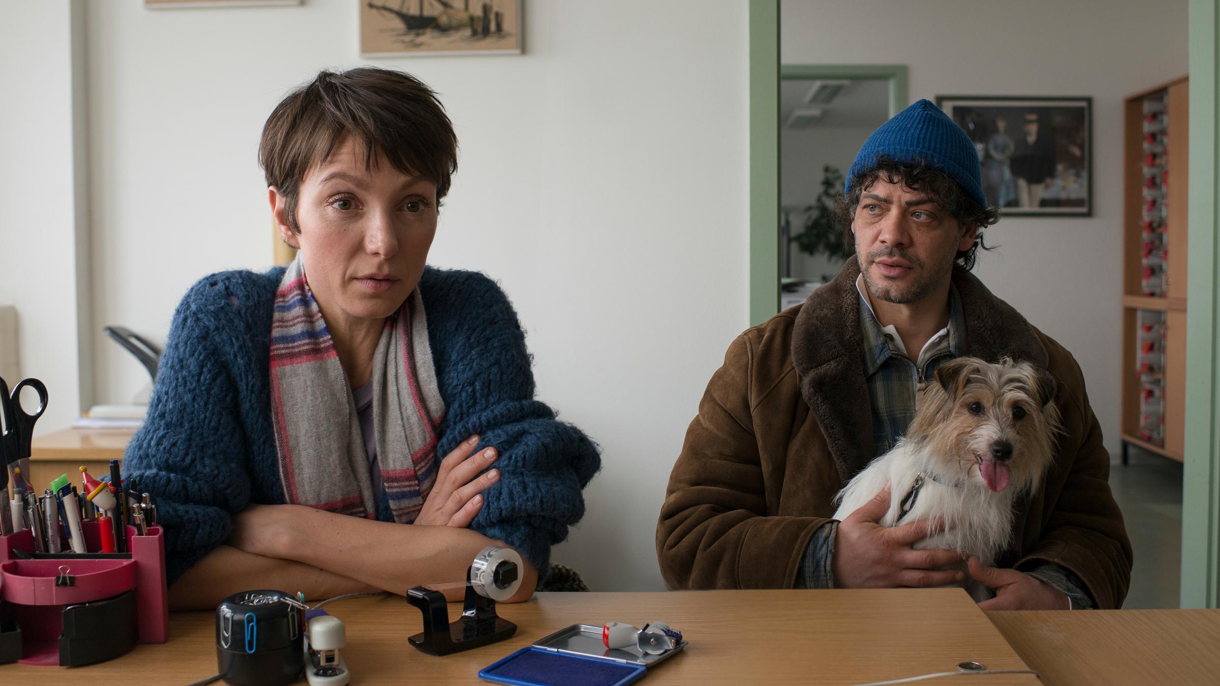 "Auf dünnem Eis": Ira (Julia Koschitz) und Konrad (Carlo Ljubek), mit Hund auf dem Schoß, sitzen in einem Büroraum vor einem Schreibtisch. Ira, mit verschränkten Armen auf den Tisch gestützt, schaut fragend ihr Gegenüber an.