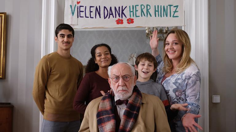 "Mein Freund, das Ekel - Voll verhintzt": Trixie Kuntze (Alwara Höfels) steht mit ihren Kindern Murat (Julius Gabriel Göze), Afia (Latisha Kohrs) und Sean (Lior Kudrjawizki) glücklich lachend und posierend beisammen. Olaf Hintz (Dieter Hallervorden) sitzt mit genervtem Blick vor den vier. Im Hintergrund hängt ein Plakat mit dem Schriftzug "Vielen Dank Herr Hintz".