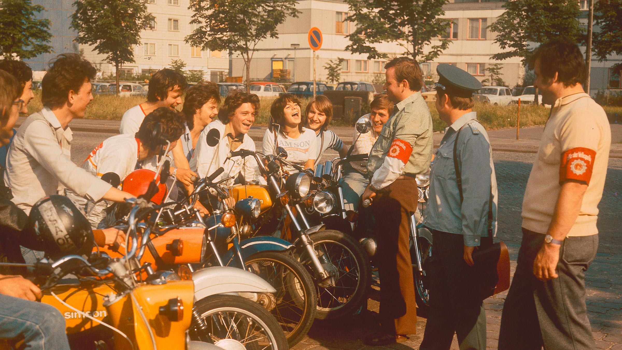 "Jugend in der DDR - Vom Stillstand zum Widerstand": Mehrere Jugendliche sitzen an einer Straße auf ihren Mopeds und sprechen mit drei DDR-Volkspolizisten.