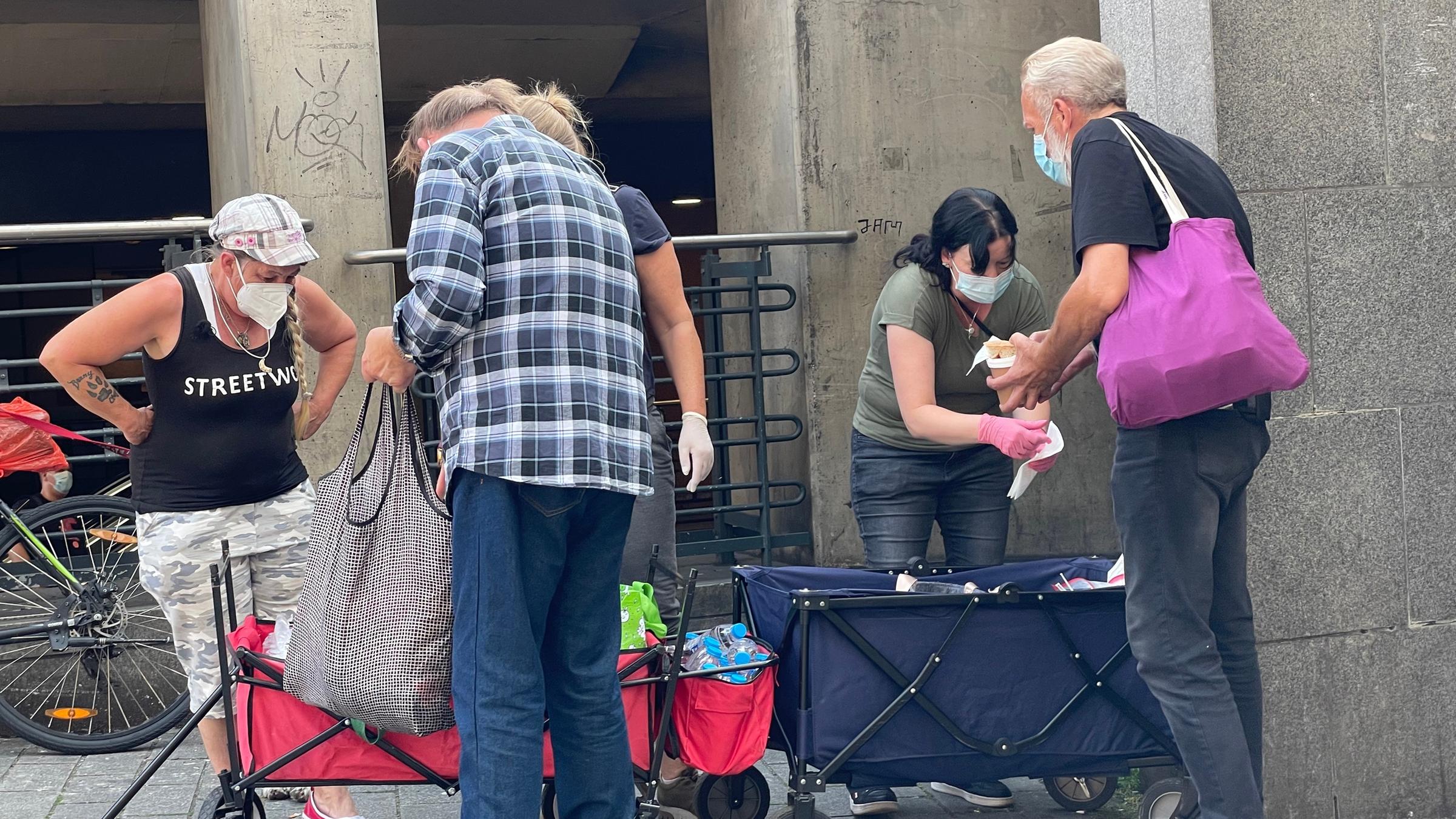"Arm gegen Arm - Verteilungskampf am unteren Rand": Vor einem Betonbau wählen zwei Menschen Hilfsgüter aus zwei mobilen Behältern aus. Weitere kümmern sich um die Vergabe, alle tragen Corona-Schutzmasken.