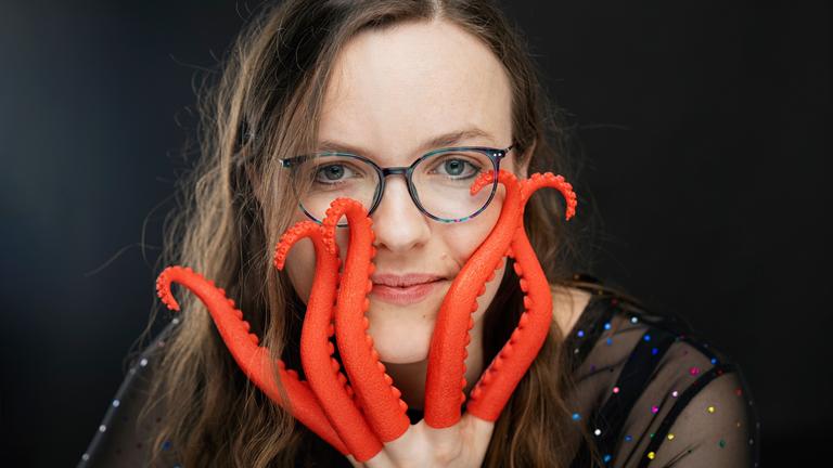 "Helene Bockhorst: Die Bekenntnisse der Hochstaplerin Helene Bockhorst": Porträt von Helene Bockhorst. Sie schaut in die Kamera und hält ihre linke Hand teilweise vor ihr Gesicht. An ihren Fingern befinden sich rote Plastik-Tintenfischarme.