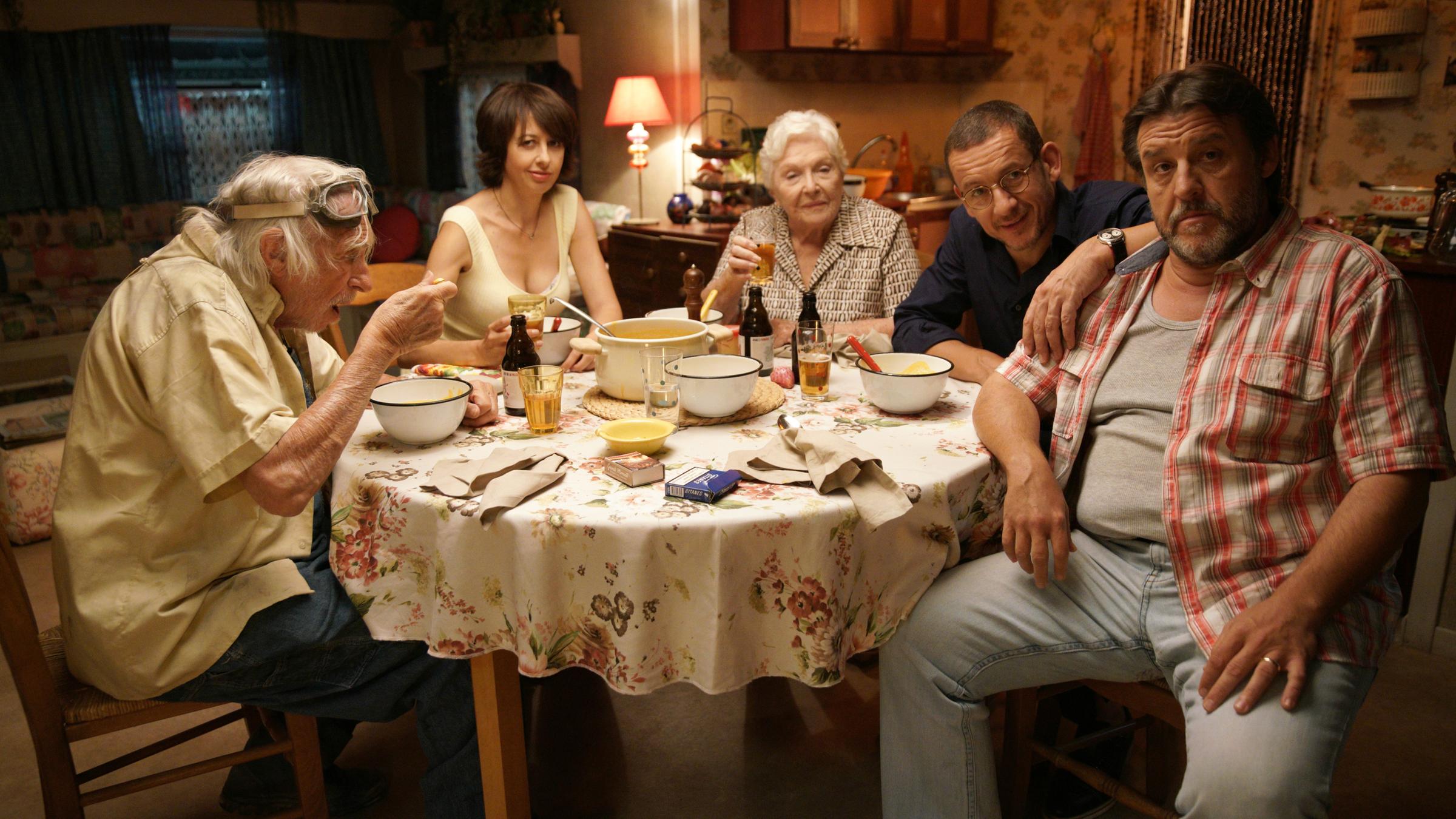 "Die Sch'tis in Paris - Eine Familie auf Abwegen": Valentin (Dany Boon), sein Vater (Pierre Richard), seine Schwägerin (Valérie Bonneton), seine Mutter (Line Renaud) und sein Bruder Gustave (Guy Lecluyse) sitzen an einem gedeckten Esstisch und schauen in die Kamera.