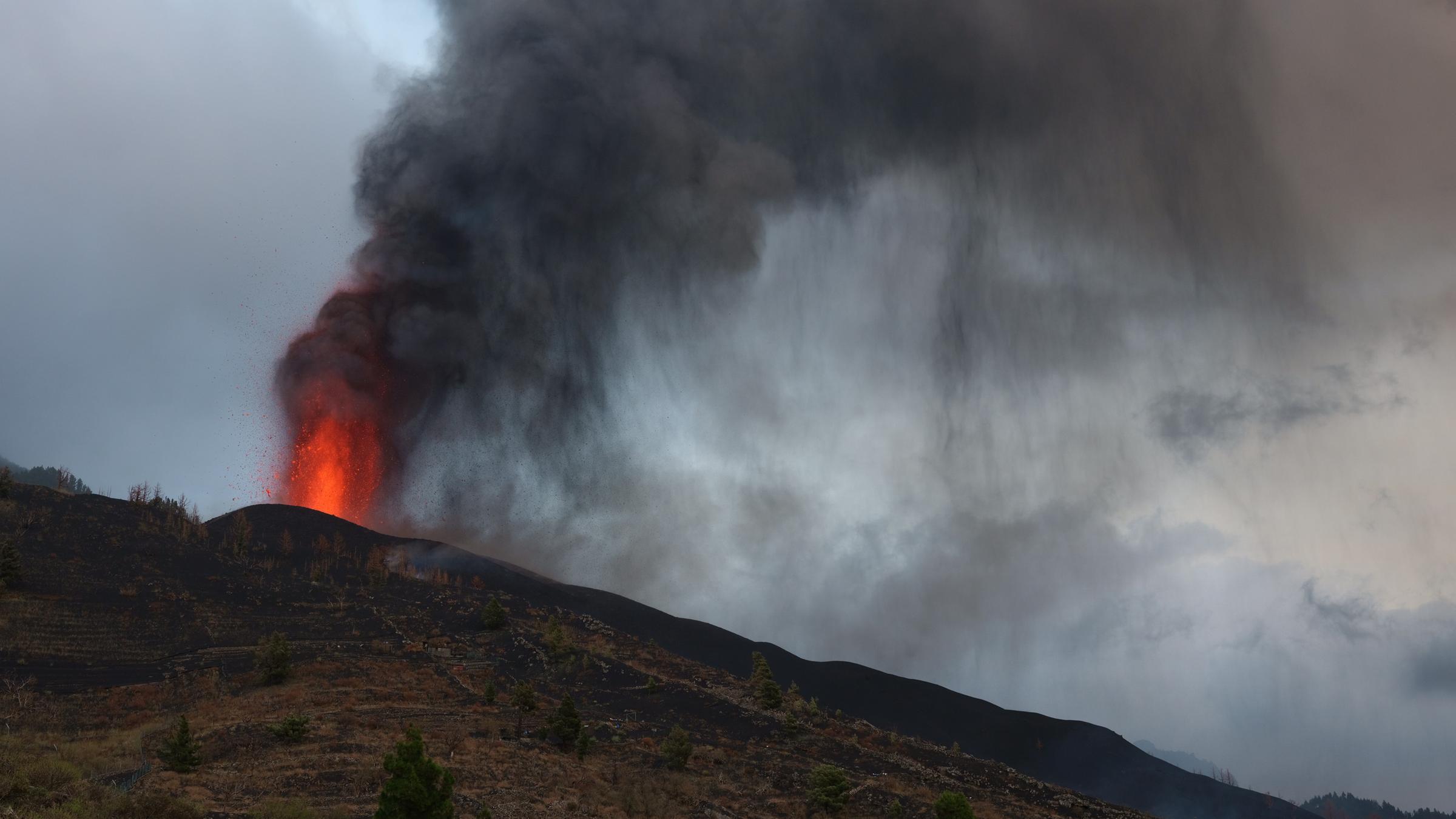 "Vulkanausbruch auf La Palma - Zerstörung und Neuanfang": Tag zwei des Vulkanausbruchs auf La Palma. Der Himmel ist grau bedeckt und eine schwarze Rußwolke liegt über dem Hang der Vulkankette Cumbre Vieja. Aus dem neu entstandenen Aschehügel kurz unter dem Grat steigt eine noch kleine Feuerfontäne auf. Es ist der neu entstandene Krater. An seinem Fuß bedeckt ein längliches dunkles Lavafeld den Berg und lässt erahnen, zu was der Vulkan noch fähig sein wird. Ein rotes Haus rechts im Vordergrund steht nur unweit entfernt von dem Fluss.