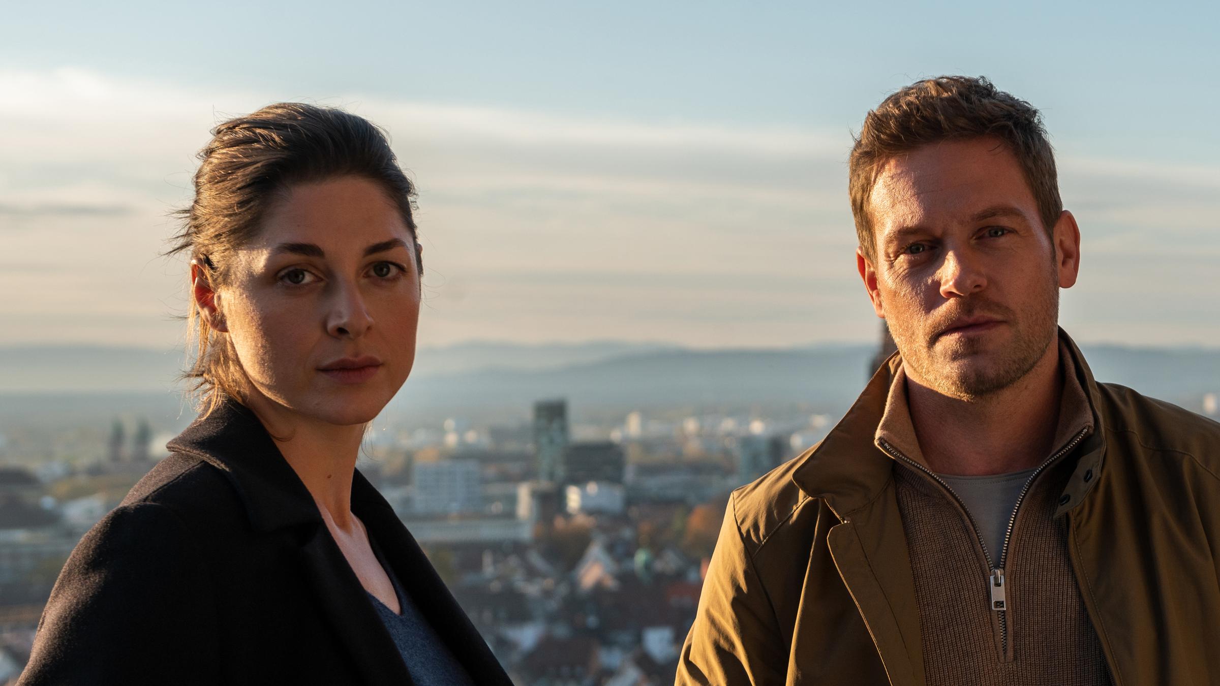"Breisgau - Nehmen und Geben": Tanja Wilken (Katharina Nesytowa) und Dennis Danzeisen (Joscha Kiefer) stehen am Geländer eines Aussichtspunkts und blicken entschlossen in die Kamera. Hinter ihnen ist das Stadtpanorama von Freiburg zu sehen.