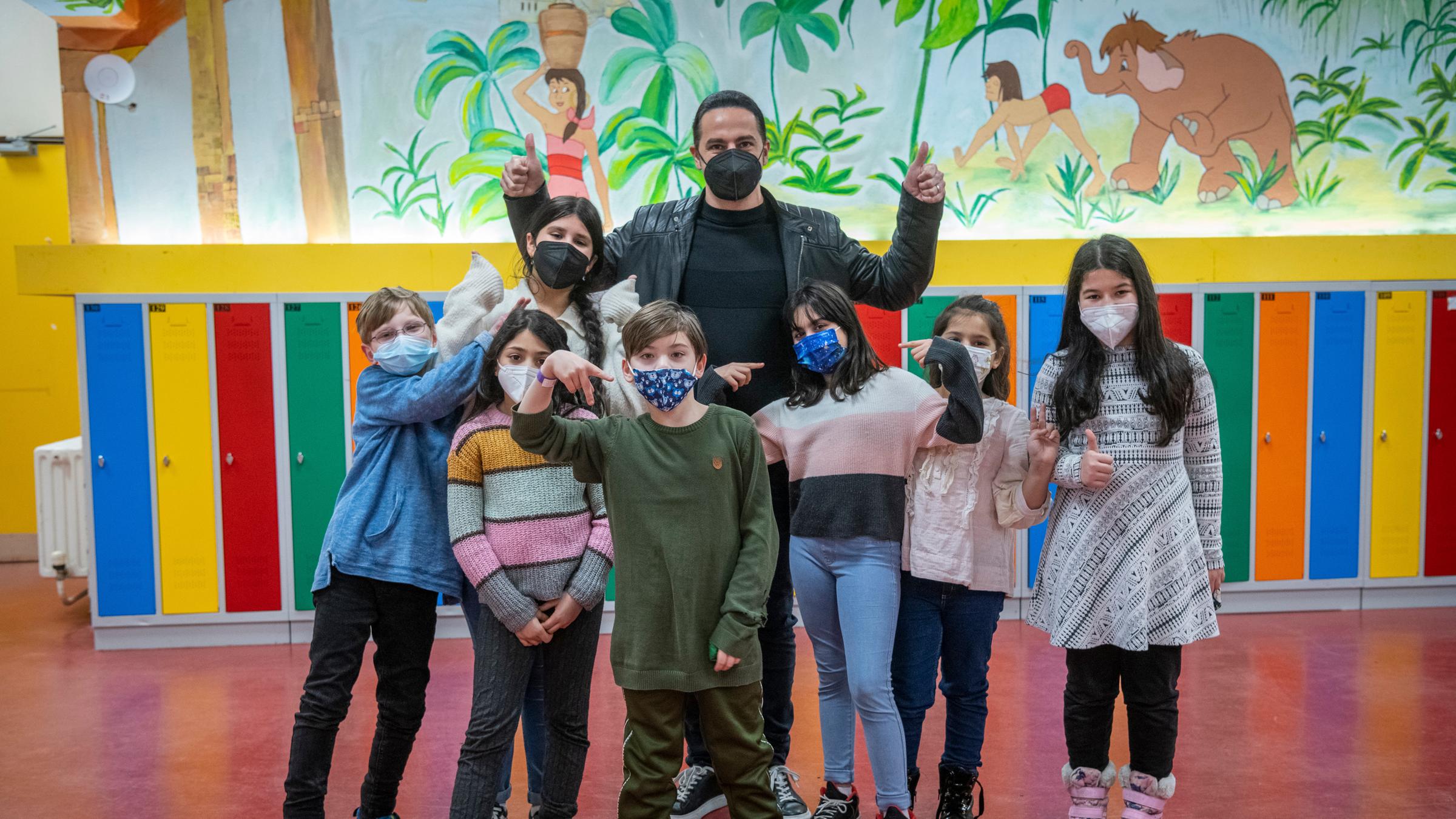 "Don't Stop the Music": In der Aula der Schule Campus Efeuweg. Bülent Ceylan posiert mit einer Gruppe von sieben Kindern. Seine Arme sind nach oben angewinkelt und seine Finger formen ein "Daumen hoch". Die Gruppe steht vor bunten Spinten. Darüber ist eine Szene aus dem Dschungelbuch mit bunten Farben gezeichnet.