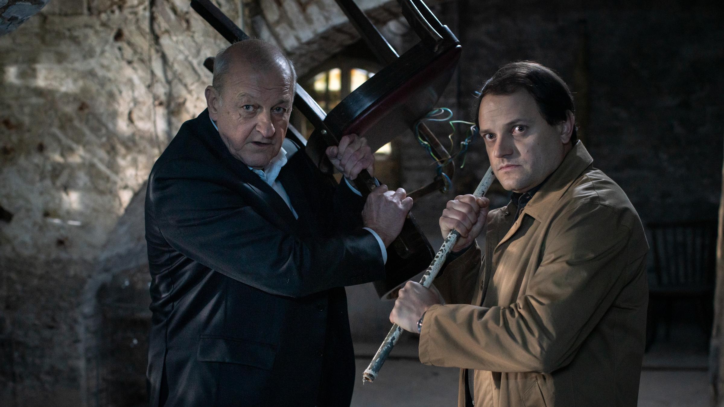 "Wilsberg - Ungebetene Gäste": Wilsberg (Leonard Lansink) und Kommissar Drechshage (Stefan Haschke) stehen im Gewölbekeller des Schlosshotels. Wilsberg hebt einen alten Stuhl hoch, Drechshage eine Eisenstange, als ob sie damit gleich zuschlagen wollten.