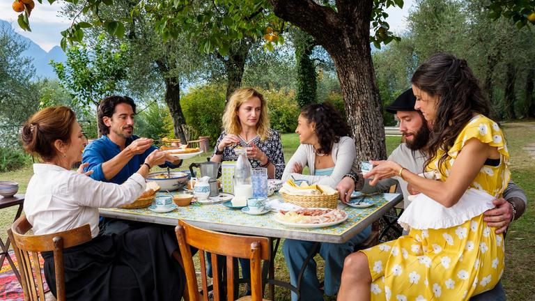 "Ein Sommer am Gardasee": Unter Orangenbäumen hat sich die Familie Ferri und Nell zum Frühstück an einem großen Tisch versammelt: Giovanna (Daniela Ziegler), Simone (Stefano Berndardin), Nell (Pia Micaela Barucki), Viola (Samirah Breuer), Diego (Sascha Pederiva) und Rosa (Liane Forestieri), die auf seinem Schoß sitzt.