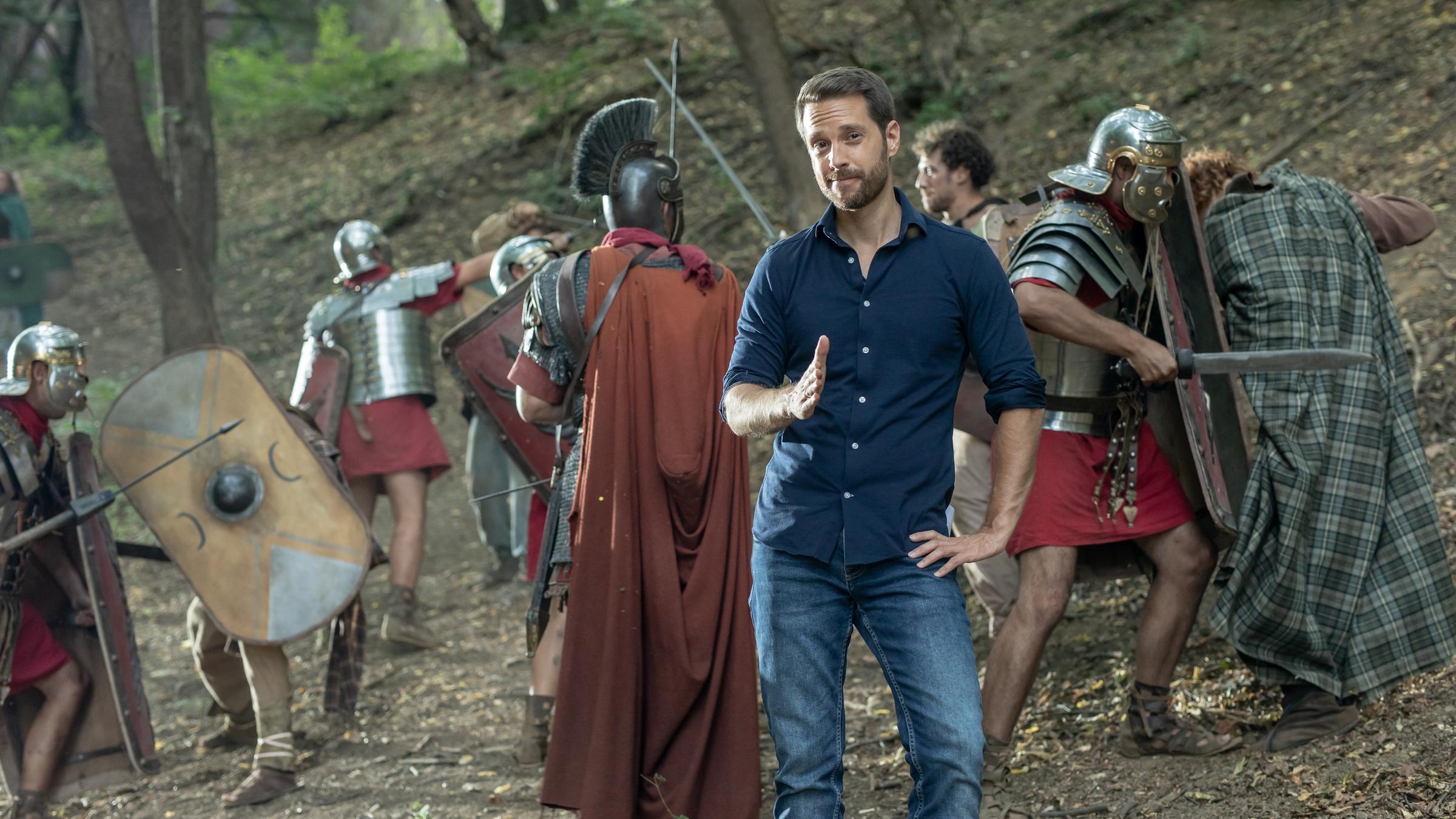 "Terra X: Ein Moment in der Geschichte - Die Varusschlacht": Spielszene: Mirko Drotschmann mit blauem Hemd und Jeans steht in einem Wald. Hinter ihm kämpfende Darsteller in Kostümen.