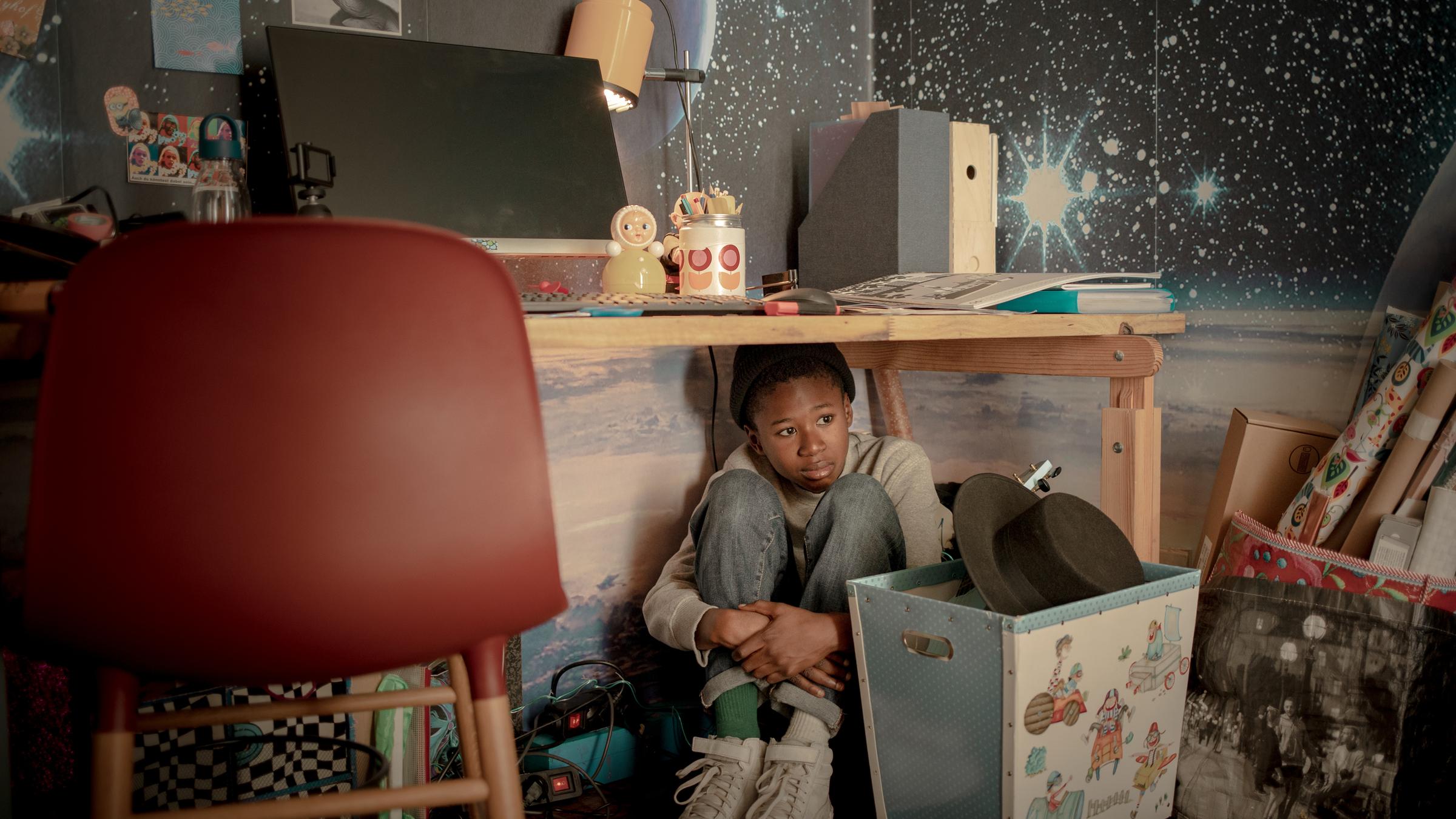 "Geheime Schatten": Nick (Aaron Sansi) kauert ängstlich und verstört schauend mit angezogenen Beinen unter dem Schreibtisch in seinem Zimmer.