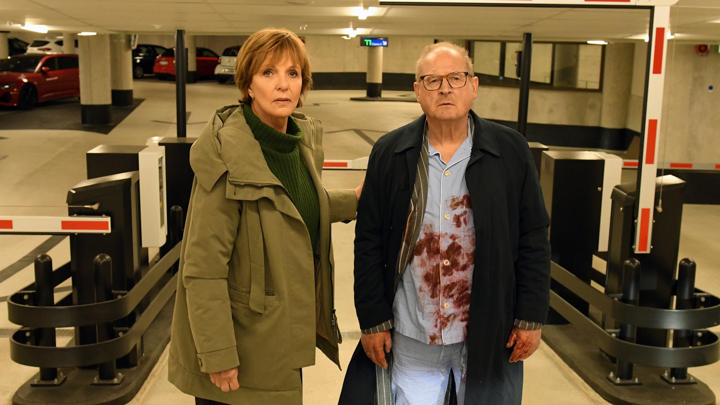 "Kommissarin Lucas - Goldrausch": Ellen Lucas (Ulrike Kriener) und Wolfgang Löhns (Burghart Klaußner) stehen nebeneinander in einem Parkhaus vor der Einfahrtsschranke. Ellen Lucas hat die Hand auf Wolfgang Löhns' Arm gelegt, der unter seinem Mantel einen blutverschmierten Schlafanzug trägt.