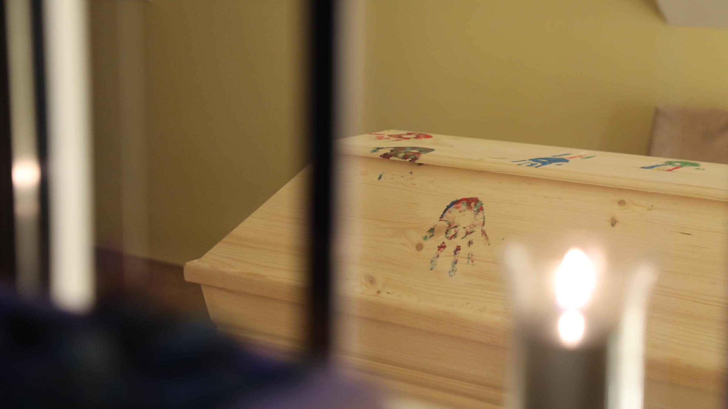 "plan b: Leben mit dem Ende - Vom anderen Umgang mit der Trauer": Ein Holzsarg ist mit bunten Handabdrücken verziert. Davor brennt eine Kerze.