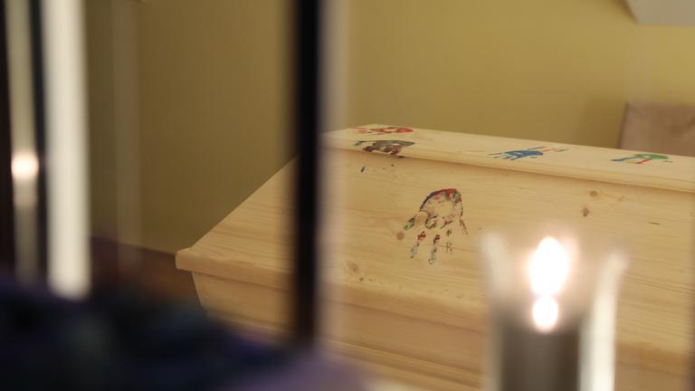"plan b: Leben mit dem Ende - Vom anderen Umgang mit der Trauer": Ein Holzsarg ist mit bunten Handabdrücken verziert. Davor brennt eine Kerze.