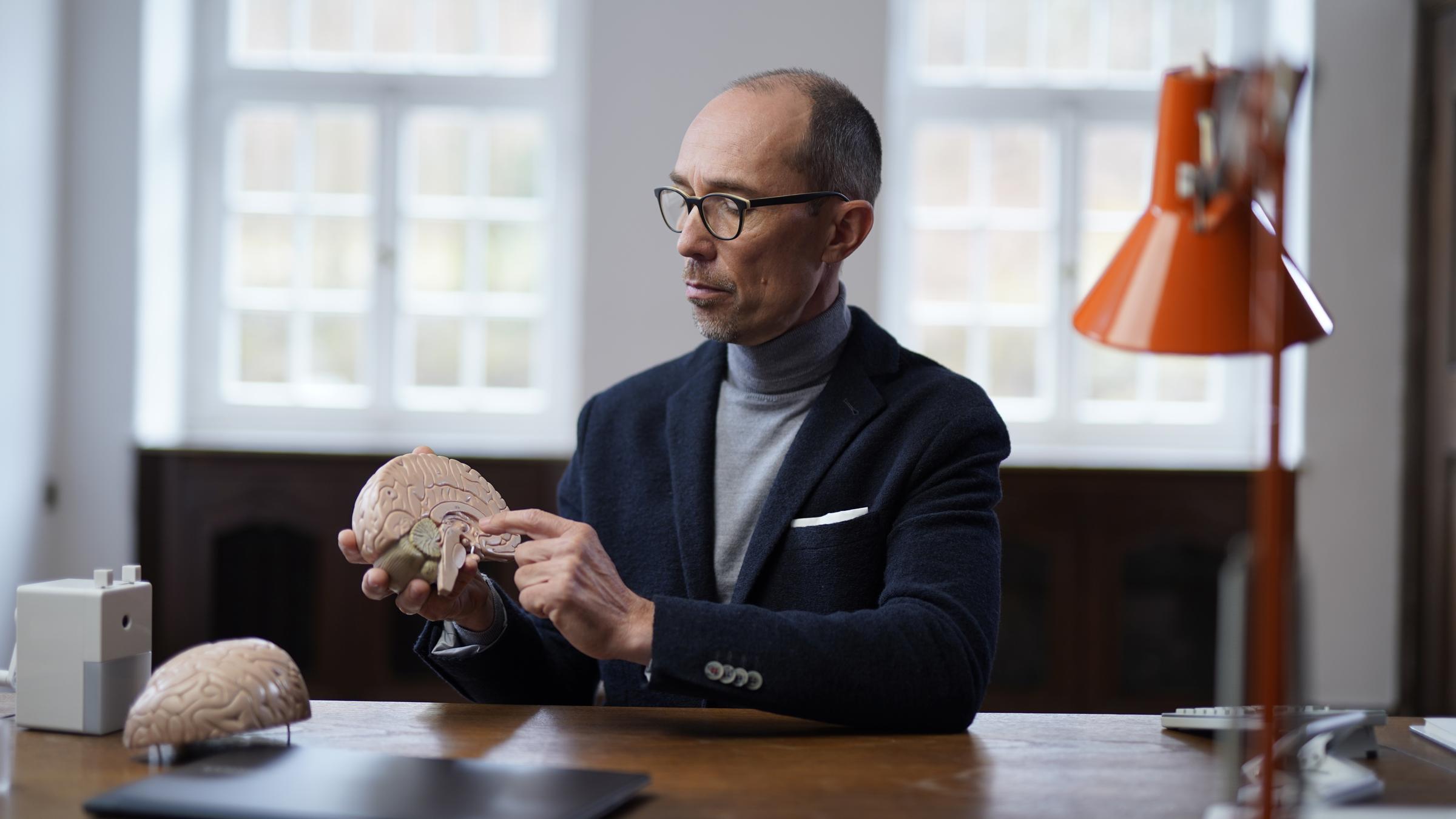 "Gesünder arbeiten, besser leben: Teilzeit für alle?": Ein Mann sitzt an einem Schreibtisch. In der Hand hält er das Modell eines Gehirns.