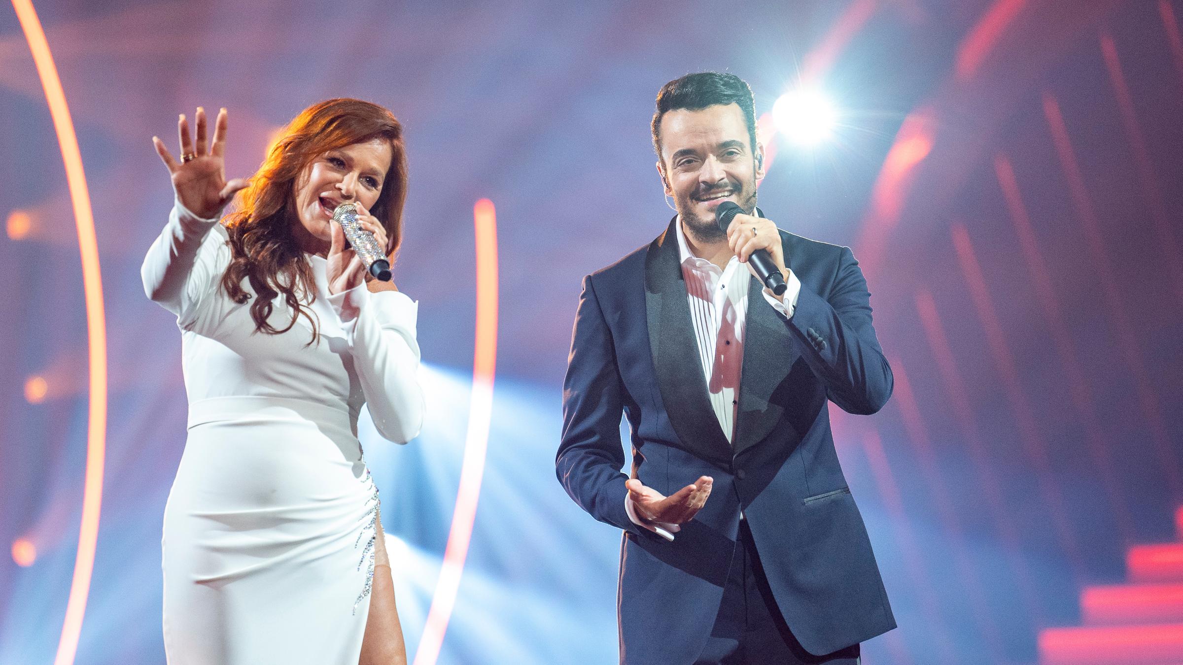 "Giovanni Zarrella präsentiert: 30 Jahre Andrea Berg": Andrea Berg im weißen Abendkleid steht neben Giovanni Zarrella im dunklen Anzug auf der Bühne. Beide haben ein Mikrofon in der Hand und singen.