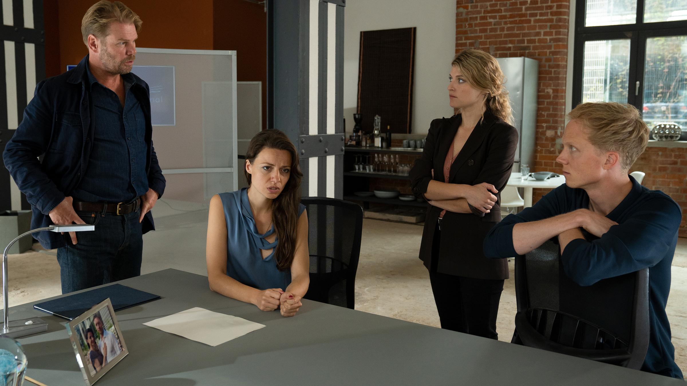 "Erzgebirgskrimi - Tödliche Abrechnung": Dr. Hellmanns Geschäftspartner Julia Jacobi (Julia Hartmann) und Phillip Mertens (Tobias Schormann) sitzen in einem großen Büro an einem Tisch. neben und hinter ihnen stehen die Kommissare Robert Winkler (Kai Scheve) und Karina Szabo (Lara Mandoki) und befragen sie.