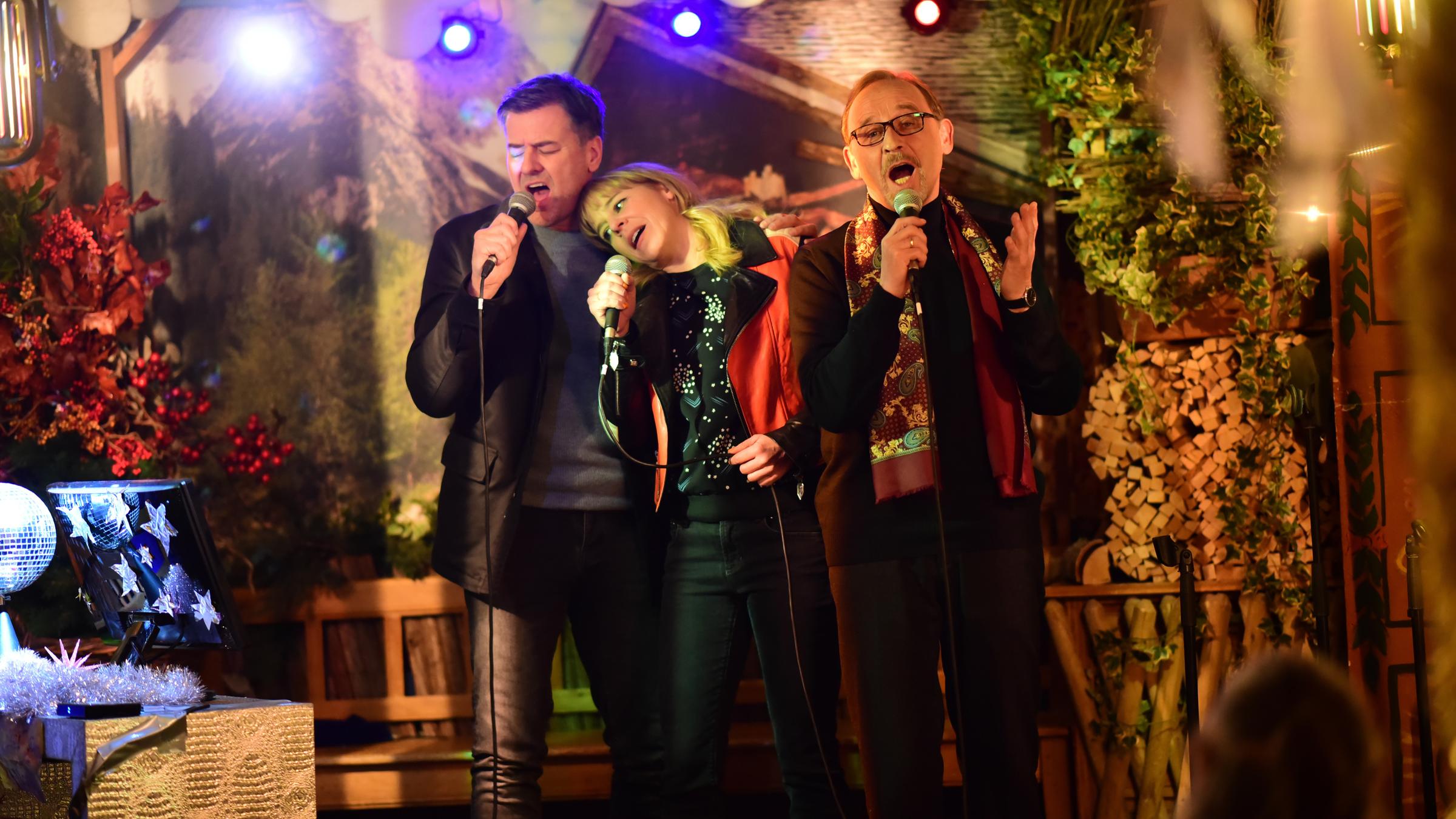 "München Mord – Schwarze Rosen": Harald Neuhauser (Marcus Mittermeier), Angelika Flierl (Bernadette Heerwagen) und Ludwig Schaller (Alexander Held) stehen jeweils mit einem Mikrofon in der Hand, zu dritt Arm in Arm, auf einer kleinen Bühne und singen.