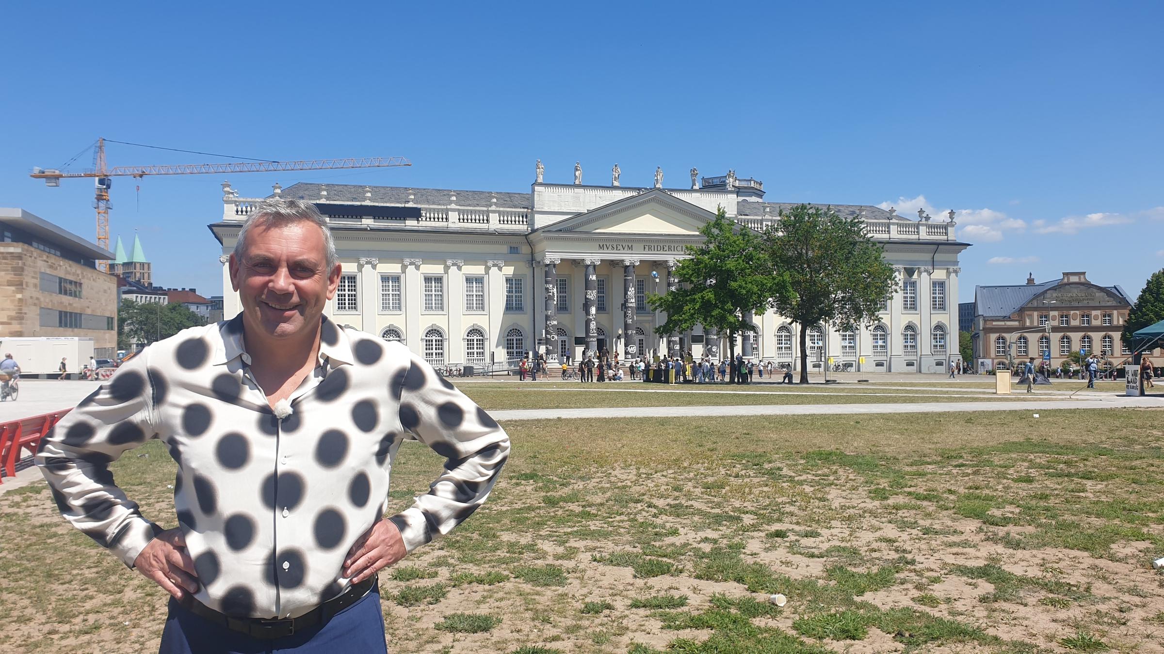 "Kaminer Inside: documenta 15" (3sat): Wladimir Kaminer steht lachend vor der Kamera, die Hände in die Hüften gestützt. Im Hintergrund sieht man das Fridericianum in Kassel. Die Sonne scheint.