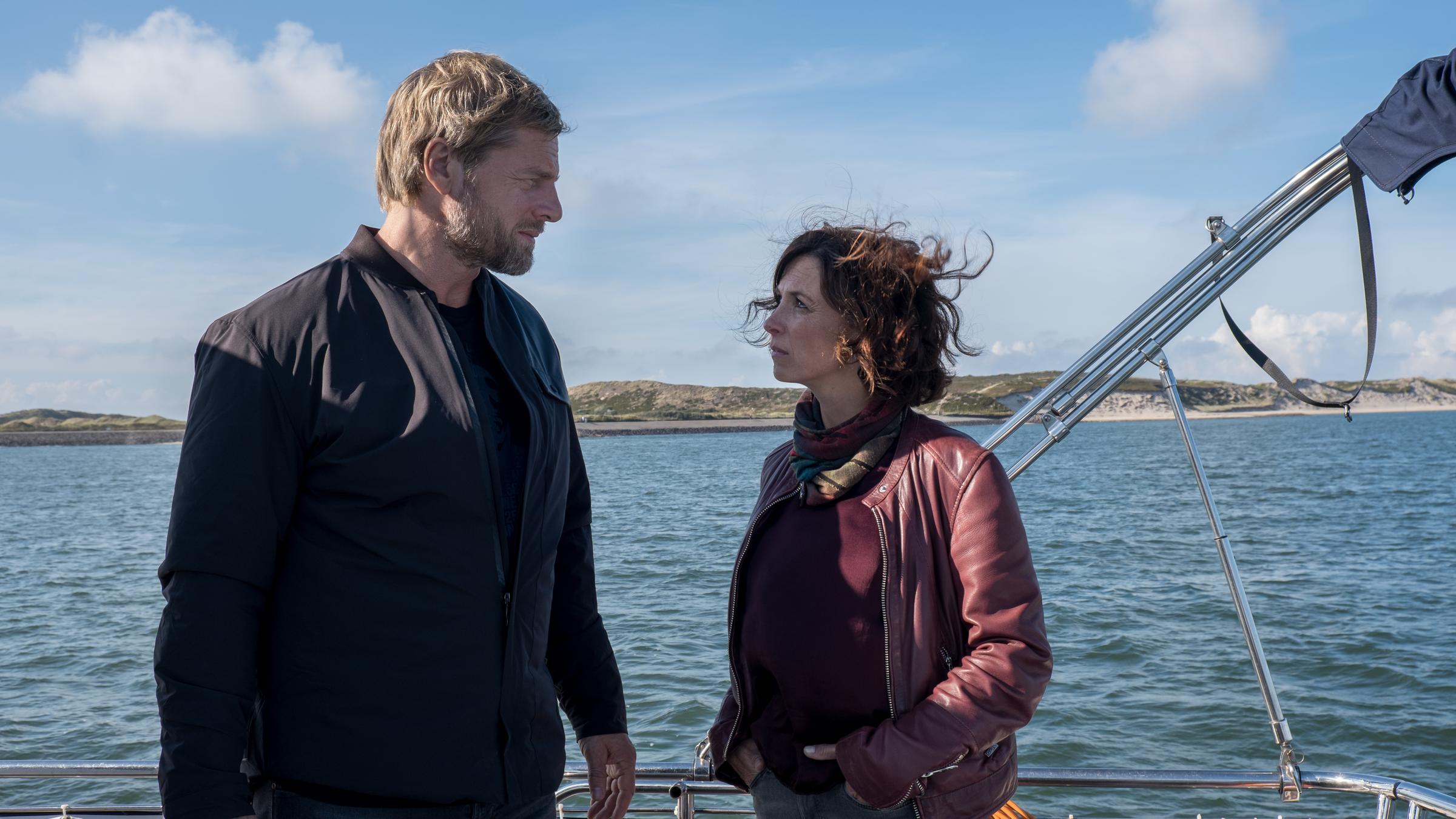 "Nord Nord Mord - Sievers sieht Gespenster": Philipp Vandamm (Henning Baum) steht Ina Behrendsen (Julia Brendler) an Deck seiner Yacht gegenüber. Hinter ihnen sieht man das offene Meer und in der Ferne die naturbelassene Küste Sylts, mit ihren Dünen und dem weißen Sandstrand. Es ist ein schöner Tag. Inas offene Haare wehen im Wind.