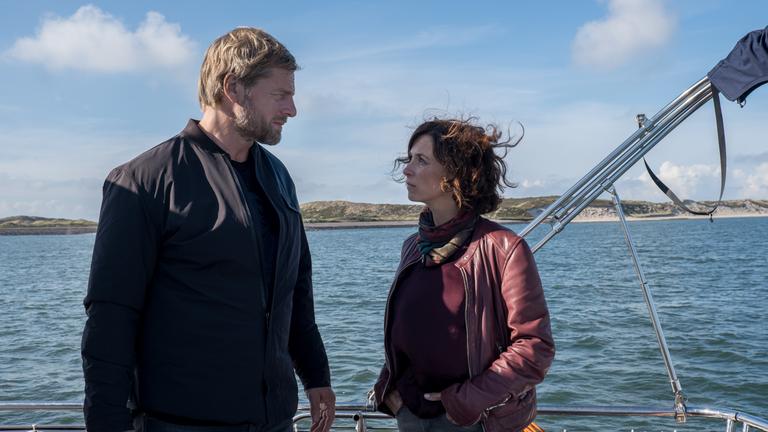 "Nord Nord Mord - Sievers sieht Gespenster": Philipp Vandamm (Henning Baum) steht Ina Behrendsen (Julia Brendler) an Deck seiner Yacht gegenüber. Hinter ihnen sieht man das offene Meer und in der Ferne die naturbelassene Küste Sylts, mit ihren Dünen und dem weißen Sandstrand. Es ist ein schöner Tag. Inas offene Haare wehen im Wind.