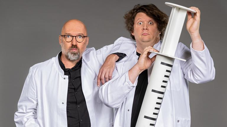 "Der satirische Jahresrückblick 2022": Die Satirekollegen Andreas Wiemers und Werner Doyé stehen in Arztkitteln nebeneinander und  blicken in die Kamera. Werner Doyé präsentiert dabei mit aufgerissenen Augen  eine überdimensionale Spritze