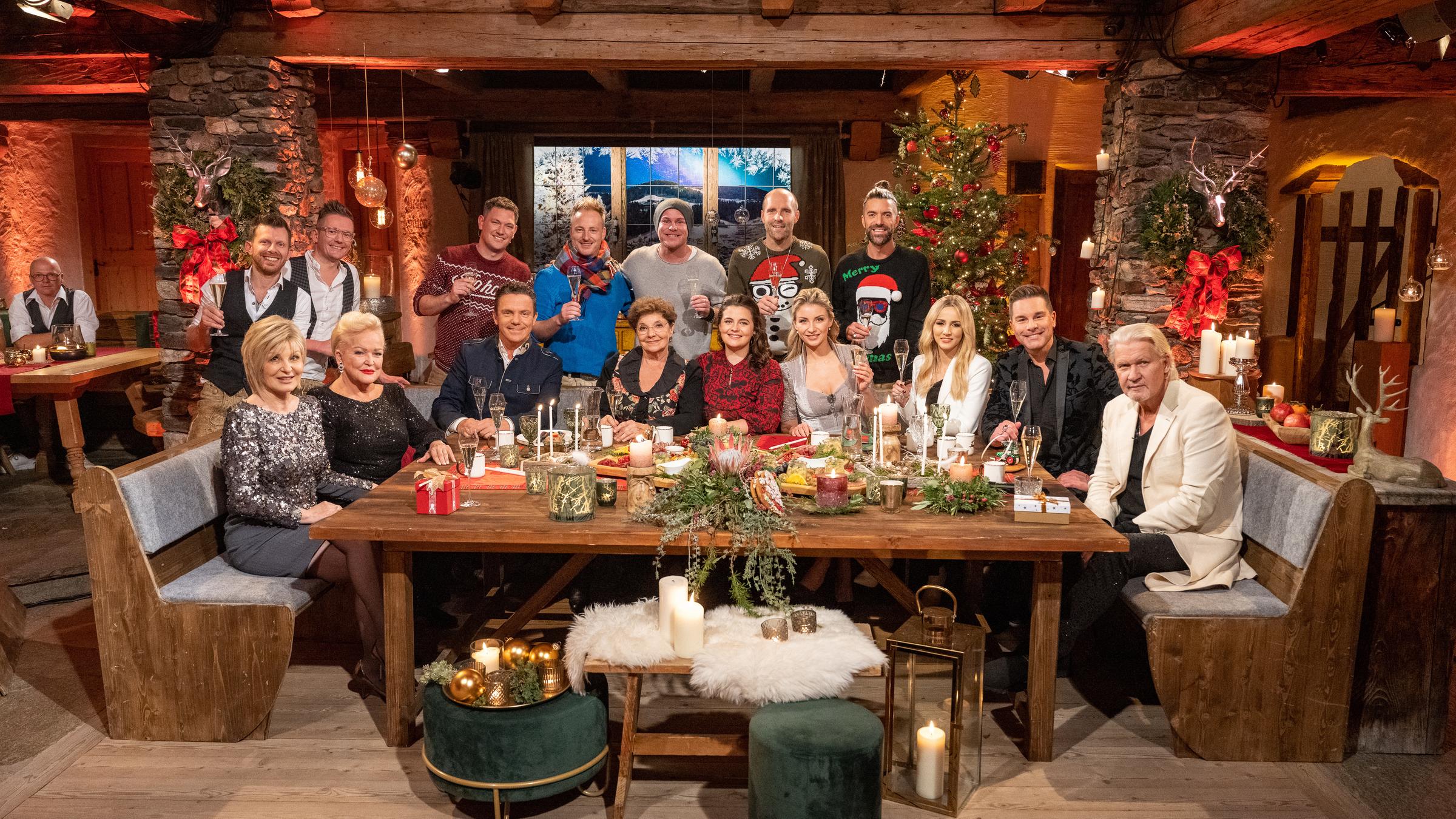 "Heiligabend mit Carmen Nebel": Carmen Nebel, Angelika Milster, Stefan Mross, Monika Baumgartner, Ronja Forcher, Anna-Carina Woitschack,  Melissa Naschenweng, Eloy de Jong und Johnny Logan vor weihnachtlicher Kulisse sitzen an einem Tisch, der ebenfalls festlich dekoriert ist. Hinter ihnen stehen Andreas Salvenmoser, Martin Schipflinger und die fünf Mitglieder der Gruppe voXXclub. Einige halten ein Glas in der Hand und lächeln in die Kamera.