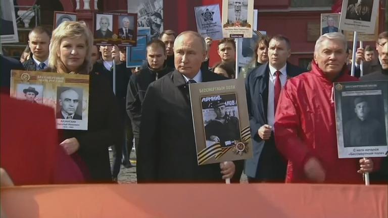 "Putins Propagandamaschine - Das manipulierte Volk": Wladimir Putin trägt ein Bild seines Vaters. Er ist umringt von weiteren Personen, die schwarz-weiße Porträts von Angehörigen tragen.