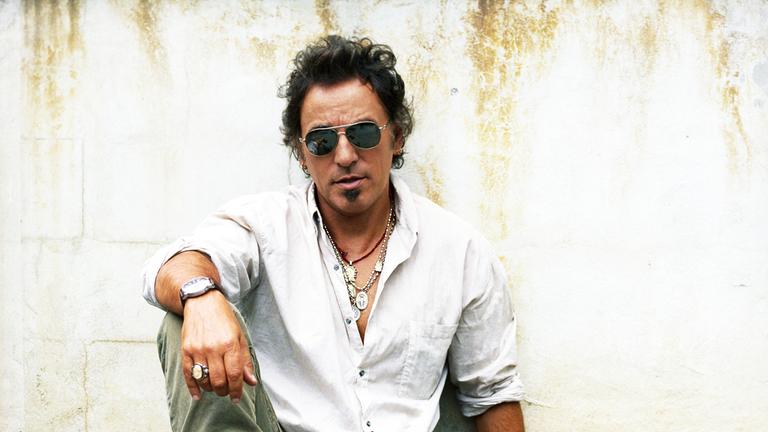 "Bruce Springsteen: Live In Barcelona": Der Sänger sitzt vor einer Mauer und blickt in die Kamera. Das rechte Bein ist angewinkelt. Der rechte Arm liegt auf dem Knie. Bekleidet ist er mit einem weißen Hemd. Er trägt einige silberne Ketten und eine dunkle Piloten-Sonnenbrille.