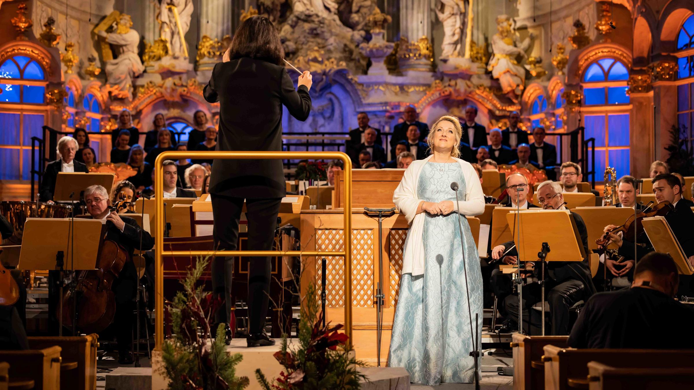 "Adventskonzert aus Dresden": Die Sopranistin Diana Damrau. Sie steht vor dem Orchester in der Frauenkirche und blickt nach oben. Um die Schultern trägt sie eine weiße Federstola. Links von ihr die Dirigentin Marie Jacquot mit dem Rücken zur Kamera. Im Hintergrund die Staatskapelle Dresden.