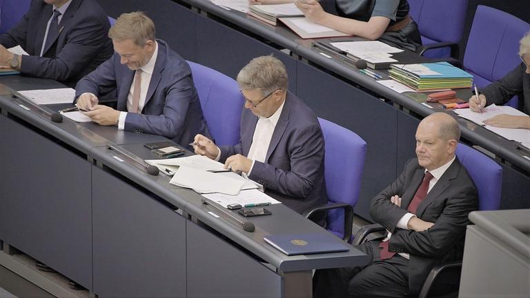 "ZDFzoom: Ampel im Ausnahmezustand": Auf der Regierungsbank sitzen Bundeskanzler Olaf Scholz, Wirtschaftsminister Robert Habeck und Finanzminister Christian Lindner nebeneinander. Scholz und Habeck schauen auf Papiere, die vor ihnen liegen, Lindner lehnt sich mit verschränkten Armen zurück.
