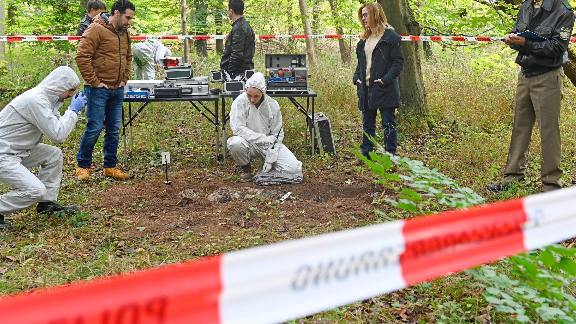 "Aktenzeichen XY... gelöst!": Beamtinnen und Beamte befinden sich tagsüber an einem abgesperrten Tatort im Wald. Die Spurensicherung untersucht die halb vergrabene Leiche.